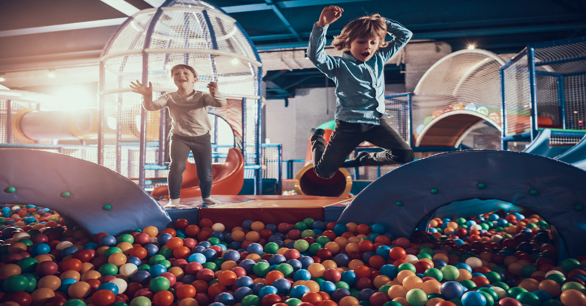 Two young boys jump from a soft play structure into a ball pit at an indoor playground.
