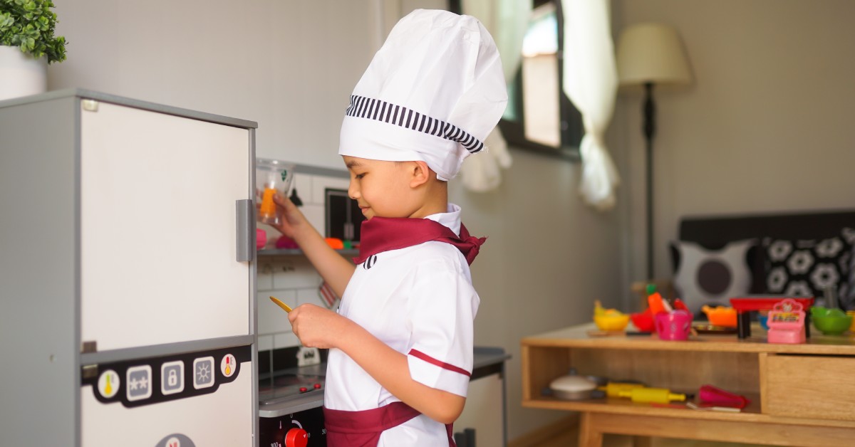 A young boy wearing a chef's costume stands in front of a toy refrigerator in his living room, with a sofa and coffee table in the background.