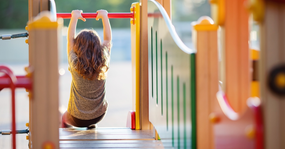 A young child swings from a bar at the end of a bridge in a colorful plastic playground structure.