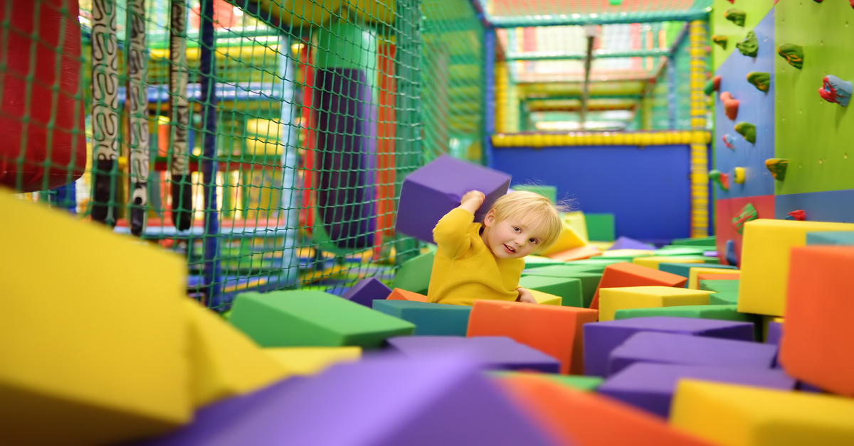 A little boy wearing a yellow sweater plays among colorful soft foam cubes in an indoor playground.